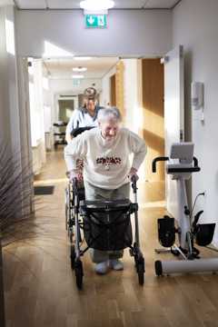 Ældre dame går ned ad en gang. Der er en SOSU-assistent, som følger efter og hjælper hende.