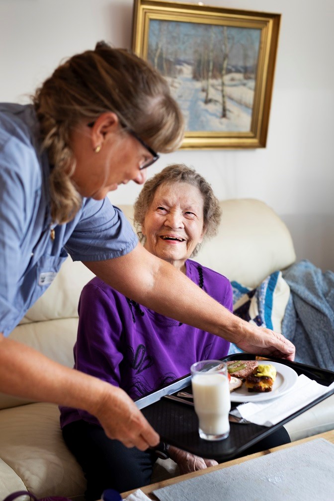 Ældre dame smiler, mens en SOSU-hjælper placerer hendes frokost foran hende.