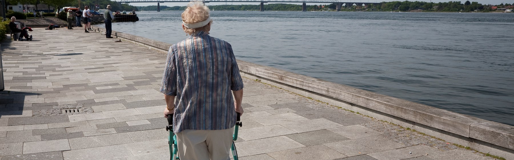 Ældre person går langs en flod. Personen støtter sig op ad sin rollator.