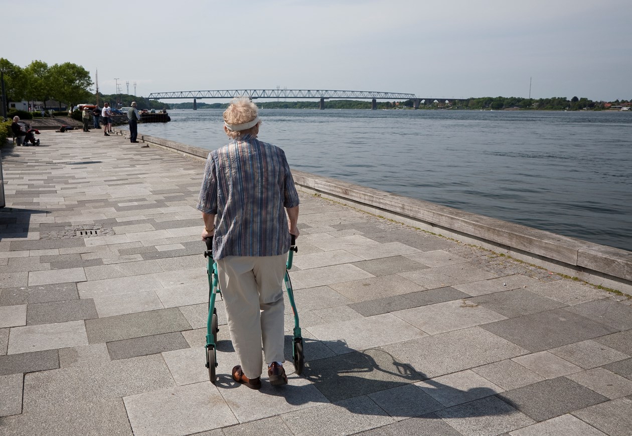 Ældre person med rollator går en tur ved vandet. Man kan se en bro i distancen.