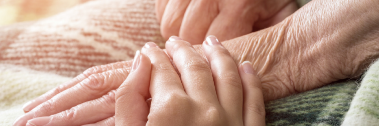 En yngre kvinde holder en ældre borger i hånden. Deres hænder ligger på et tæppe på den ældre persons skød.