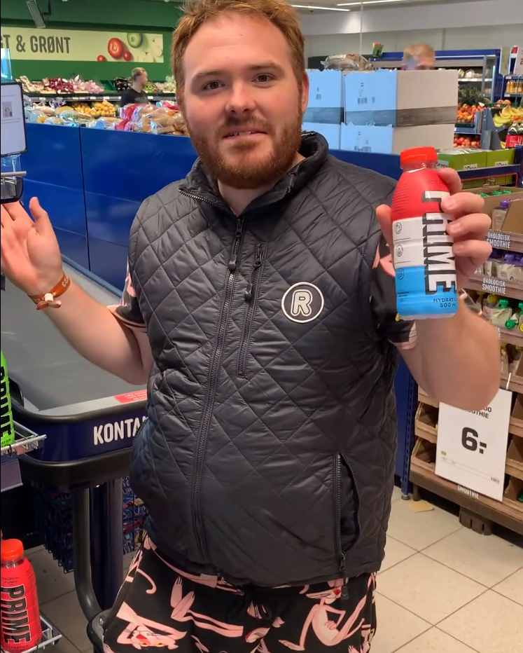 Mand med Rema 1000-vest på. Han står foran kassen og ser glad ud.