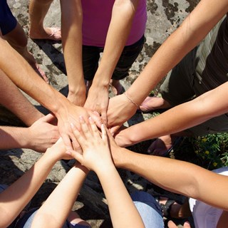 En masse personer står i en cirkel og har deres hænder samlet på midten. Det symboliserer, at de samarbejder og hjælper hinanden.